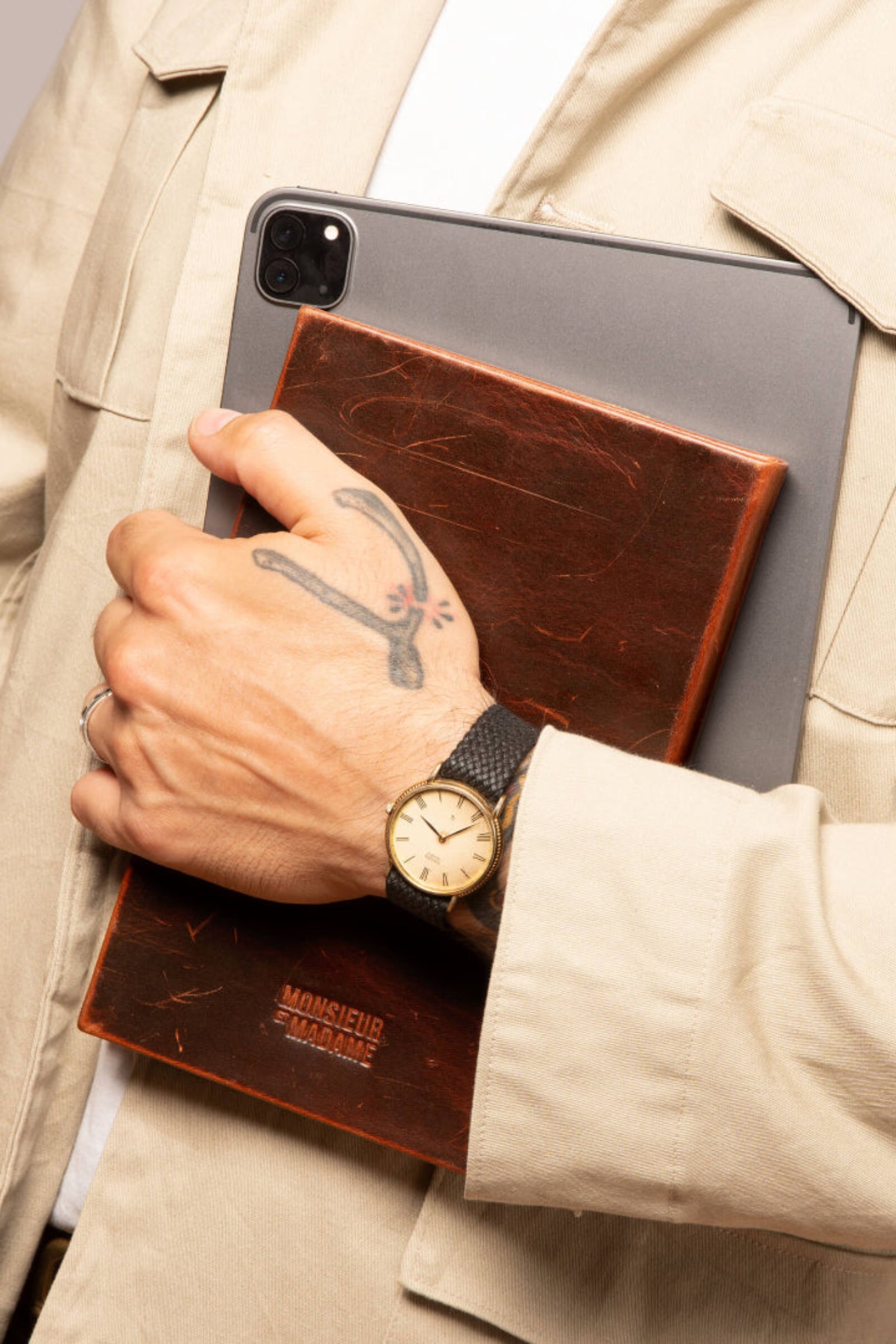 Person holding a tablet with a leather case, wearing a watch, against a neutral background